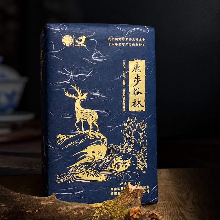 A dark blue tea or gift package with gold foil artwork of a deer, clouds, and trees, standing on a wooden surface against a dark blurred background.