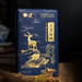 A dark blue tea or gift package with gold foil artwork of a deer, clouds, and trees, standing on a wooden surface against a dark blurred background.