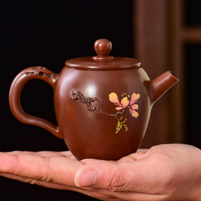 A small brown ceramic teapot with a floral decoration, held in a person's hand against a dark background.