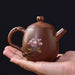 A small brown ceramic teapot with a pink floral design, held in a person's hand against a dark background.