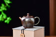 A small ceramic teapot with a textured, metallic bronze-brown glaze sits on a light beige box against a dark background with soft green foliage on the left.