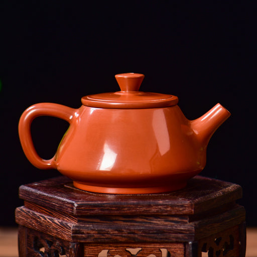 An red-orange glazed ceramic teapot with a matching lid, displayed on a dark wooden stand against a dark background with soft green foliage on the left.