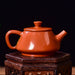 An red-orange glazed ceramic teapot with a matching lid, displayed on a dark wooden stand against a dark background with soft green foliage on the left.