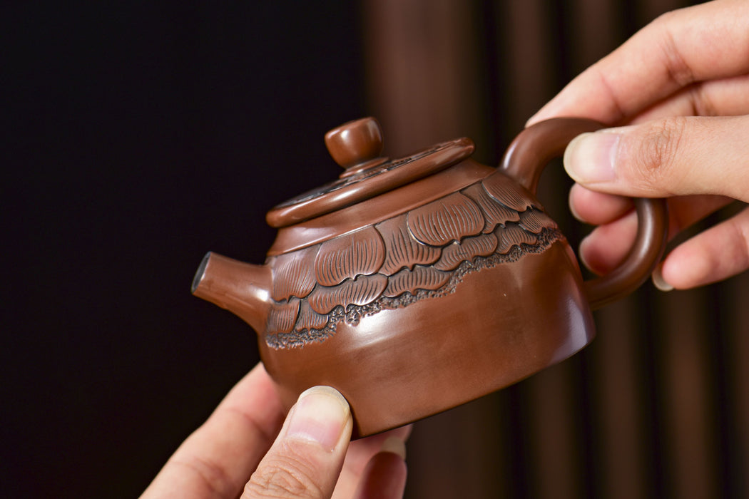 A close-up of a brown ceramic teapot with carved floral details, held by two hands against a dark, blurred background.