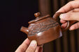 A close-up of a brown ceramic teapot with carved floral details, held by two hands against a dark, blurred background.