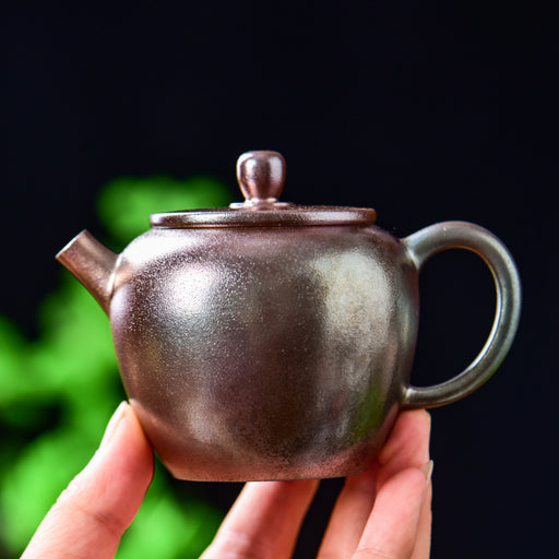A hand holding a small handmade ceramic teapot with a dark metallic bronze finish, set against a dark background with soft green bokeh.