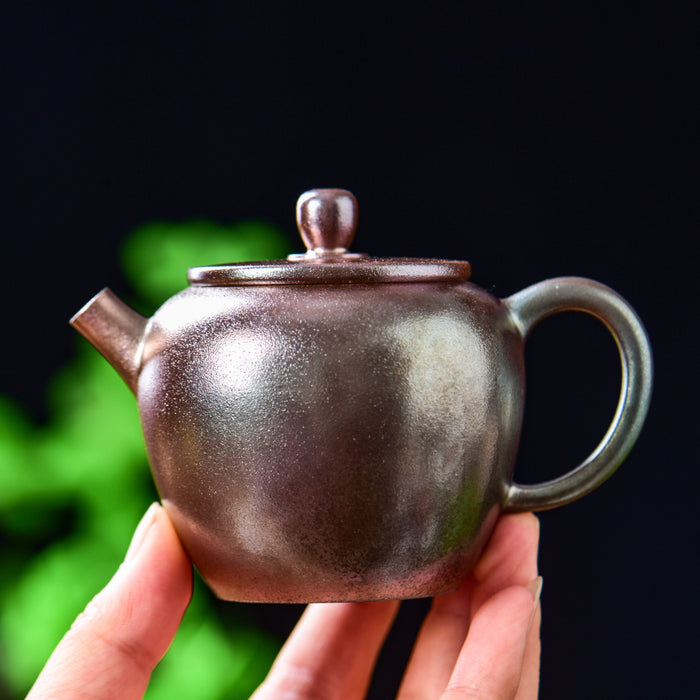 A hand holding a small handmade ceramic teapot with a dark metallic bronze finish, set against a dark background with soft green bokeh.