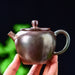 A hand holding a small handmade ceramic teapot with a dark metallic bronze finish, set against a dark background with soft green bokeh.