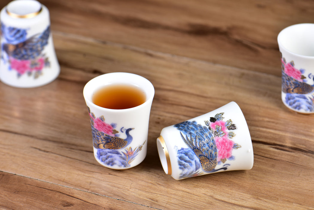 Decorative ceramic cups with floral and peacock designs on a wooden surface, one cup contains a dark liquid.