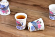 Decorative ceramic cups with floral and peacock designs on a wooden surface, one cup contains a dark liquid.
