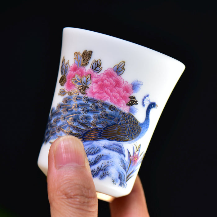 Hand holding a small ceramic cup with peacock and floral design against a dark background
