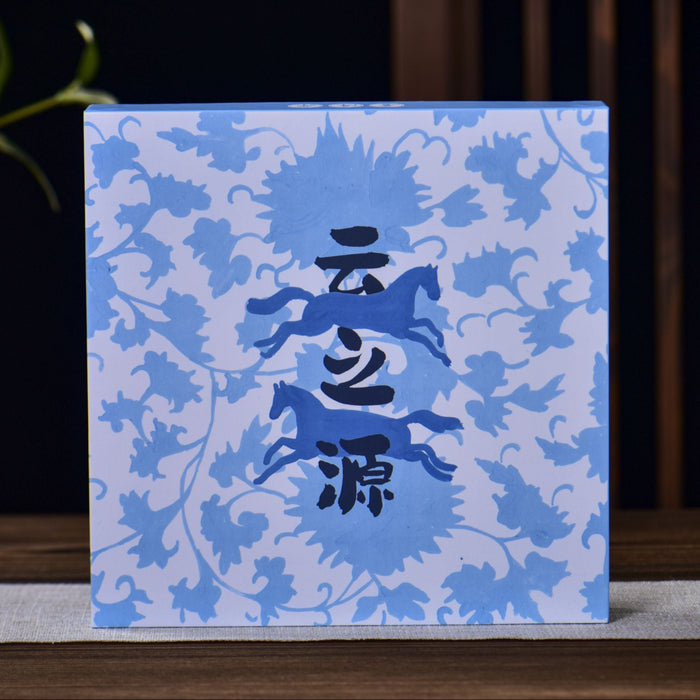 A square blue and white patterned box with black Chinese characters in the center, displayed on a wooden surface with a small ceramic pitcher and plant in the background.