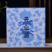 A square blue and white patterned box with black Chinese characters in the center, displayed on a wooden surface with a small ceramic pitcher and plant in the background.