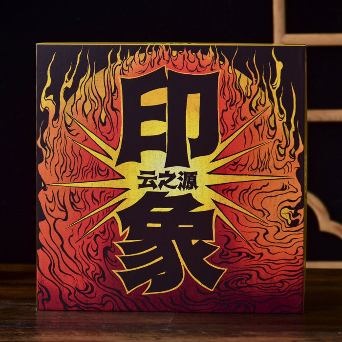 A square decorative gift box for pu-erh tea cakesstanding on a wooden surface, featuring bold black Chinese characters over a fiery red, orange, and yellow flame-like pattern. The box is photographed in a styled interior setting with a dark background, a wooden lattice screen, a teapot on the left, and a small vase on the right.
