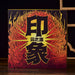 A square decorative gift box for pu-erh tea cakesstanding on a wooden surface, featuring bold black Chinese characters over a fiery red, orange, and yellow flame-like pattern. The box is photographed in a styled interior setting with a dark background, a wooden lattice screen, a teapot on the left, and a small vase on the right.