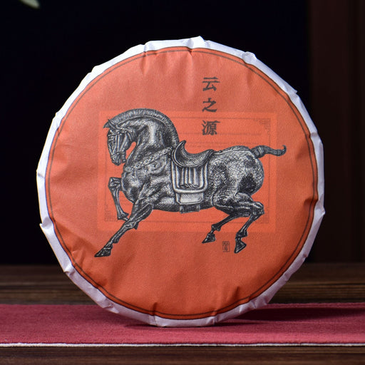 A round cake of Pu Er tea wrapped in an orange paper sleeve featuring a black horse illustration and Chinese characters, displayed on a wooden table with a teapot and ceramic vessel in a dark, traditional setting.