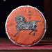 A round cake of Pu Er tea wrapped in an orange paper sleeve featuring a black horse illustration and Chinese characters, displayed on a wooden table with a teapot and ceramic vessel in a dark, traditional setting.