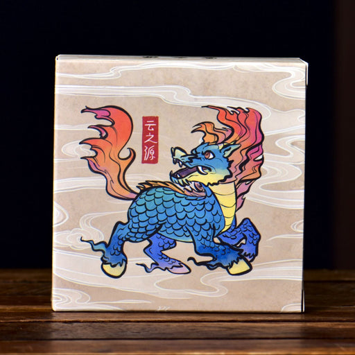 A square box containing ripe pu-erh tea with a light wood-grain background featuring a colorful illustration of a blue dragon with orange and red flames, standing on a dark wooden surface.