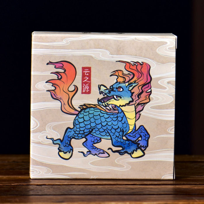A square box containing ripe pu-erh tea with a light wood-grain background featuring a colorful illustration of a blue dragon with orange and red flames, standing on a dark wooden surface.