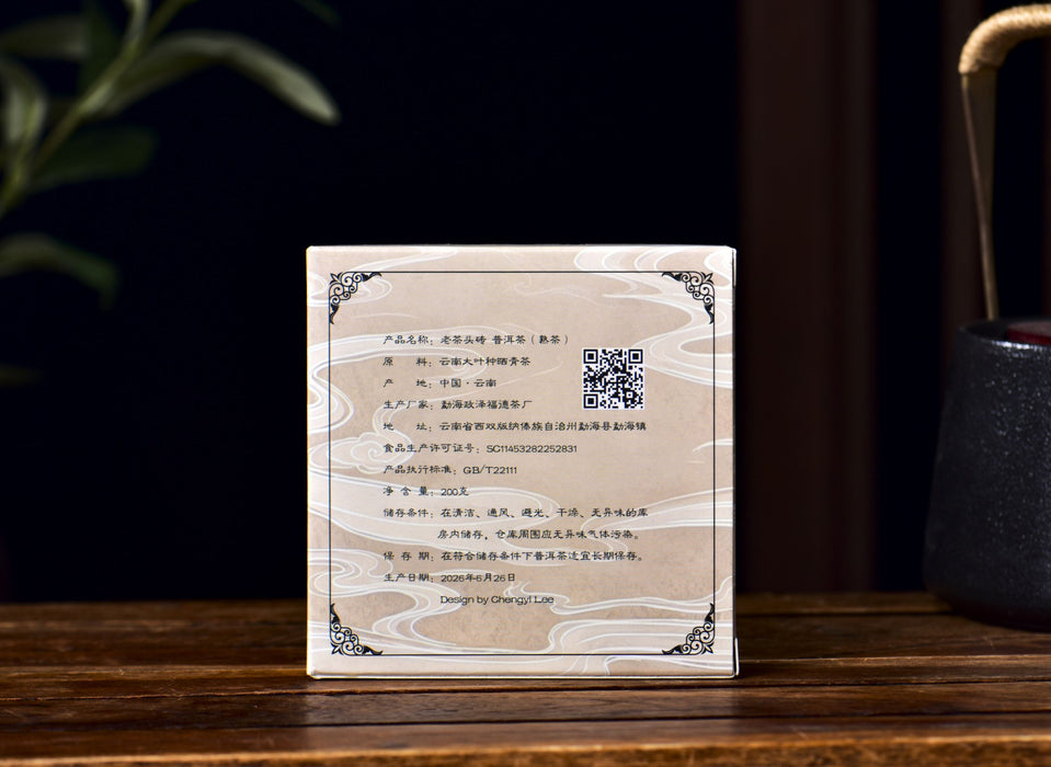 A square box containing ripe pu-erh tea cha tou with Chinese text and a QR code, featuring a cream and beige wood-grain pattern with black corner ornaments, displayed on a dark wooden table.