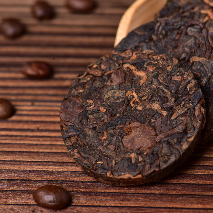 Dark, compressed tea cakes with visible leaf texture arranged in a wooden scoop on a brown wooden surface, with scattered coffee beans around them.