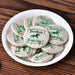 A white ceramic plate filled with round, beige fabric-wrapped mini tea cakes printed with green text reading 'Yunnan Sourcing' and a horse logo, arranged on a brown wooden surface.