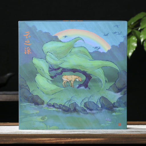 A square tea cake box featuring a stylized landscape painting with green mountains, a small deer, birds, and a rainbow, displayed on a wooden surface against a dark background with a teapot and green leaves.