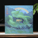 A square tea cake box featuring a stylized landscape painting with green mountains, a small deer, birds, and a rainbow, displayed on a wooden surface against a dark background with a teapot and green leaves.