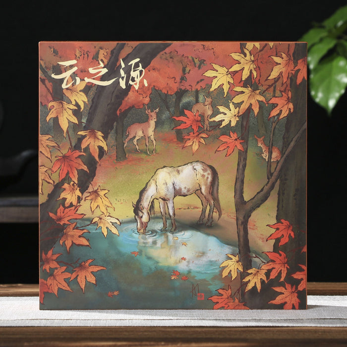 A square pu-erh tea gift box featuring a painted autumn forest scene with red and orange leaves, a white horse drinking water, and Chinese calligraphy at the top, displayed on a tabletop with a dark background and green leaves in the side.