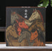 A square decorative pu-erh tea gift box or book cover featuring an illustration of several horses in warm earthy tones, standing on a tabletop with a dark background and blurred green leaves behind it.