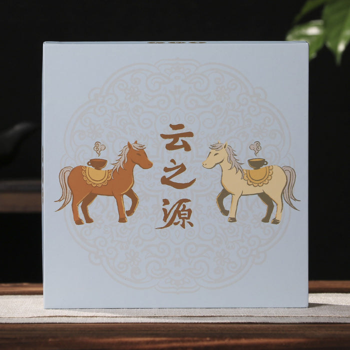 A square light blue pu-erh tea gift box box with gold Chinese characters and illustrations of two horses stands on a wooden surface against a dark background with a teapot and green leaves.