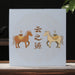 A square light blue pu-erh tea gift box box with gold Chinese characters and illustrations of two horses stands on a wooden surface against a dark background with a teapot and green leaves.