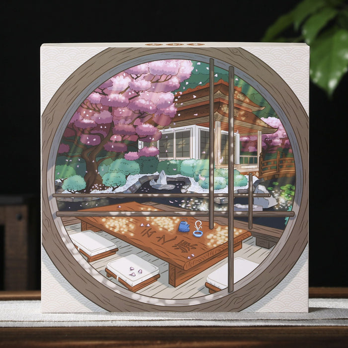 A square pu-erh tea gift box featuring a circular printed illustration of a traditional Chinese garden with cherry blossoms, displayed on a wooden surface against a dark background with a teapot and green leaves.