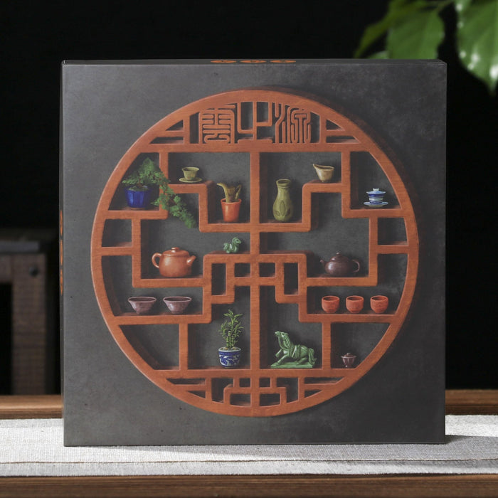 A square dark gray pu-erh tea gift box featuring a circular reddish-brown lattice design filled with small colorful ceramic tea and plant motifs, displayed on a tabletop against a dark background with a plant and teapot visible.