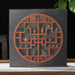 A square dark gray pu-erh tea gift box featuring a circular reddish-brown lattice design filled with small colorful ceramic tea and plant motifs, displayed on a tabletop against a dark background with a plant and teapot visible.