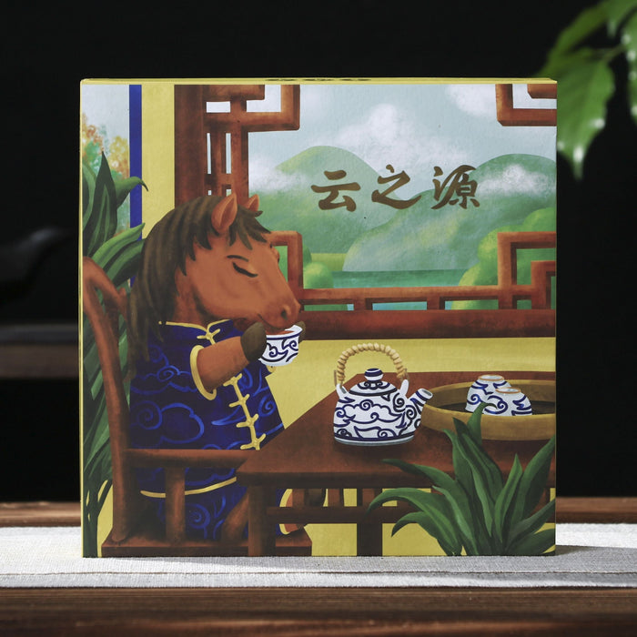 A square illustrated pu-erh tea gift box  standing upright on a tabletop, featuring a traditional Chinese scene with a horse-like figure at a table, tea vessels, greenery, and the Chinese characters '云之源'. The background is dark with a teapot on the left and green leaves on the right.