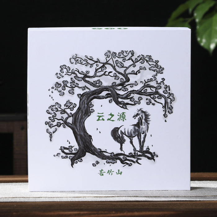 A square white pu-erh tea gift box with a black line-art illustration of a tree and horse, standing on a tabletop against a dark background with a teapot on the left and green leaves on the right.