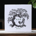 A square white pu-erh tea gift box with a black line-art illustration of a tree and horse, standing on a tabletop against a dark background with a teapot on the left and green leaves on the right.