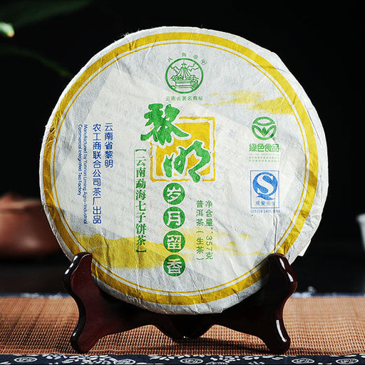 A cake-shaped package of Liming 'Everlasting Aroma' Raw Pu-erh Tea, with Chinese characters and English text on the label, displayed on a wooden stand with tea-related items in the background.
